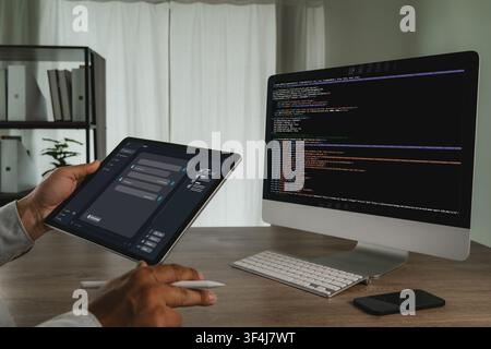 Hands hold a tablet showing an AI chatbot interface beside a desktop computer with code on screen, illustrating automated customer support and digital Stock Photo