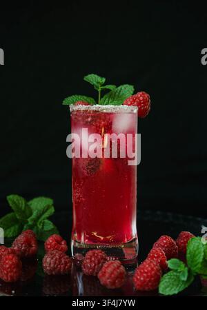 Fresh raspberry cocktail with ice cubes, mint leaves and ripe raspberries served in a tall glass against a black background. Refreshing summer beverag Stock Photo