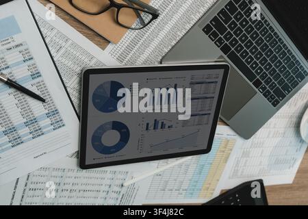 Tablet displaying investment charts beside laptop, printed spreadsheets, calculator, eyeglasses on desk, supporting market trend analysis, financial r Stock Photo