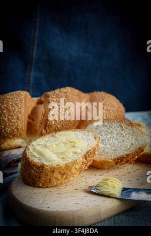 Fresh sliced white bread spread with butter on a wooden cutting board with artisan bread and rolls in the background. Rustic bakery concept. Stock Photo