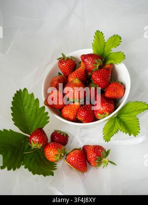 Fresh ripe strawberries in a white bowl with green leaves on a light fabric background. Organic summer berries and healthy seasonal fruit. Stock Photo