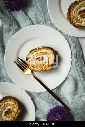 Slice of homemade sponge cake roll with cream filling served on a white plate with a dessert fork and purple flowers. Stock Photo