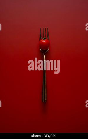A single fresh cherry tomato impaled on a metal fork against a minimalist red background. Stock Photo