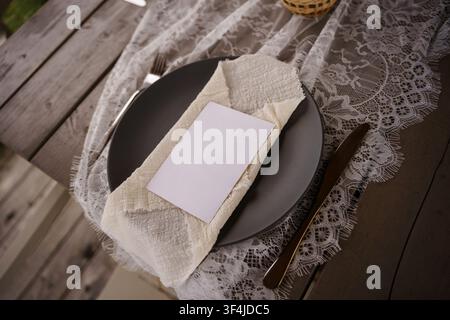 Blank paper card on dark plate with intricate lace runner, perfect for wedding menu or event stationery display on wooden table surface, table setting Stock Photo