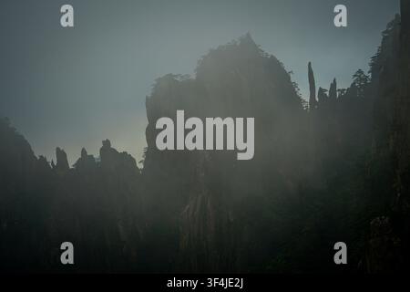 Dark granite mountain peaks rise vertically, partially shrouded in light gray fog against a muted blue-gray sky. Sparse dark green pine trees cling to Stock Photo