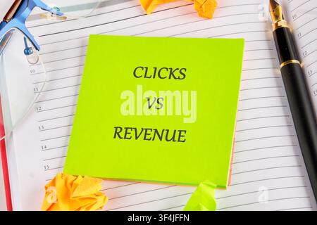 A bright green sticky note with the words clicks vs revenue is placed on a notebook in an office. Glasses and a pen are visible nearby. Stock Photo