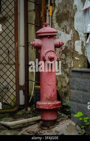 A heavily weathered red fire hydrant with peeling paint and rust stands centered in the frame, flanked by an aged, textured wall with peeling plaster Stock Photo