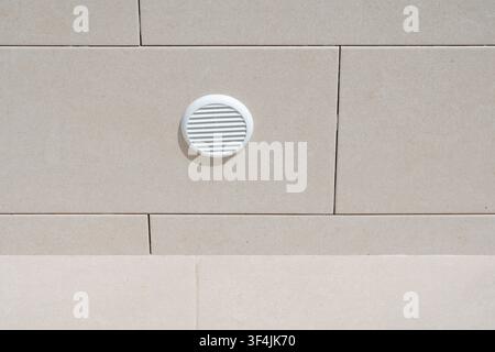 A circular white vent with horizontal slats is mounted on a textured beige stone facade, suggesting architectural detail Stock Photo