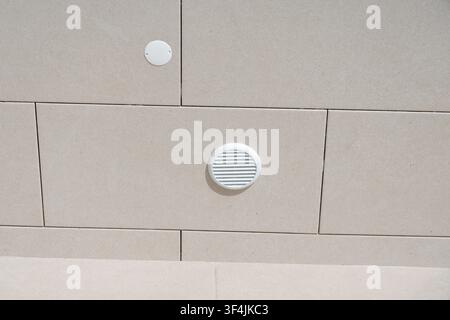 A textured beige stone wall features two circular white vents, one with slats and the other a simple cover, set against the neutral facade Stock Photo