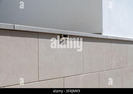 A detailed view of a modern exterior wall featuring beige tiles and a grey concrete ledge with a small metal fixture attached Stock Photo