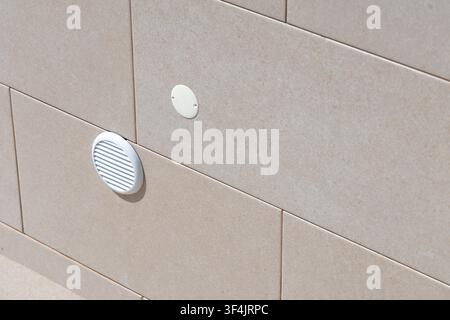 Close-up view of a light-colored tiled exterior wall featuring two small vents and a discreet light fixture Stock Photo