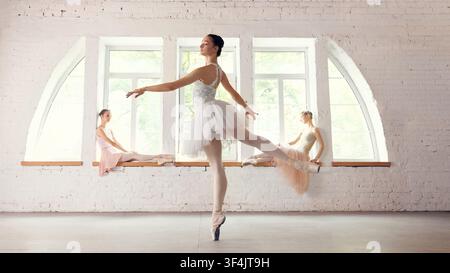 Ballet dancer balancing in foreground while group rests near windows Stock Photo