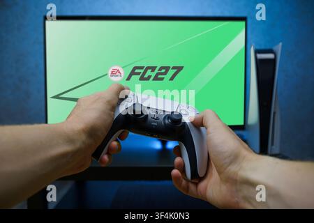 Hands holding PlayStation 5 DualSense controller in front of TV screen showing EA Sports FC 27 game, console gaming and football video game concept. Stock Photo