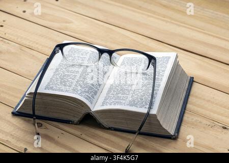 Open dictionary book with black framed reading glasses on pages resting on wooden table, concept of nearsighted vision, education and study Stock Photo