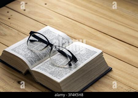 Black eyeglasses on open dictionary book on wooden table, symbolizing reading, education, study and myopia in a warm rustic setting. Stock Photo