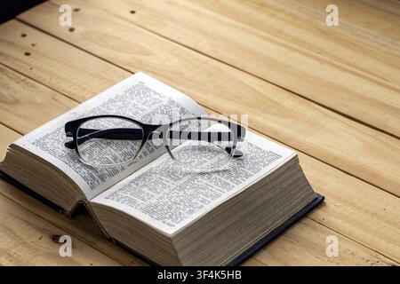 Black reading glasses on open vintage dictionary book pages resting on rustic wooden table, symbolizing nearsighted vision, reading and knowledge Stock Photo