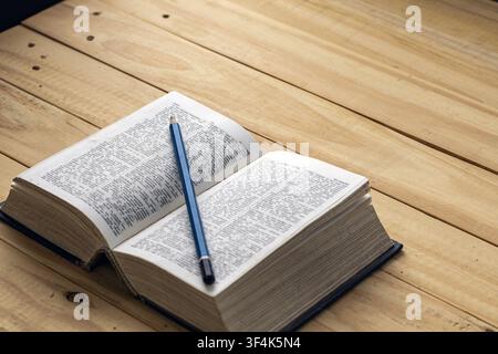 Open vintage dictionary book with sharpened blue pencil resting on printed pages, on rustic wooden table, symbolizing reading and study. Stock Photo