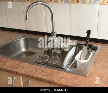 Kitchen sink features a stainless steel basin with a drying rack containing glasses and plates, alongside a knife holder on a textured countertop Stock Photo