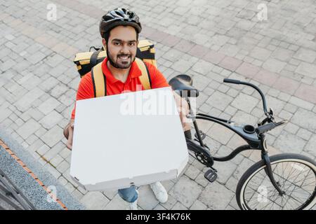 Order delivery. Smiling man with beard, in helmet, shows box of pizza to peephole Stock Photo