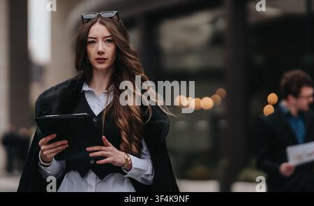 Business woman walking on city street using tablet while looking confidently at camera Stock Photo