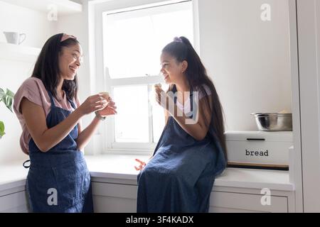 African American mom and teenage daughter sharing cupcakes in home kitchen, wearing denim aprons Stock Photo
