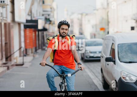Online delivery service at home. Friendly courier in helmet with backpack on bicycle rides down the street Stock Photo
