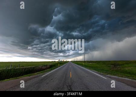 View of a long straight road stretching into the distance under a dramatic and heavy dark storm cloud formation over the green landscape in Oklahoma C Stock Photo