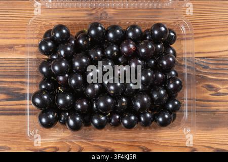 Fresh jabuticaba fruits densely packed inside a transparent plastic clamshell container on wooden planks Stock Photo
