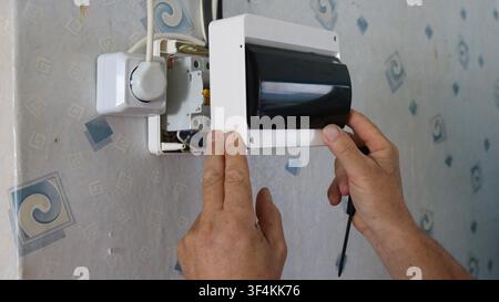 installing an electrical panel on a wallpapered wall. Hands position the white plastic housing with a dark protective cover over the circuit breakers Stock Photo