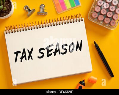 Notebook displaying TAX SEASON surrounded by office supplies, calculator, and sticky notes on a vibrant yellow background, indicating financial prepar Stock Photo