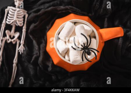 Top view of orange pumpkin mug filled with marshmallows and black spider beside skeleton decoration on textured black fabric Halloween background. Stock Photo