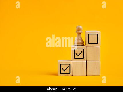 Wooden cubes with checkmark icons and a chess pawn representing task completion and step by step progress tracking on a yellow background Stock Photo
