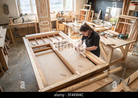 Craftswoman repairs and renovates vintage wooden window frame on workbench in woodworking shop. Stock Photo