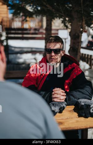 Man in red jacket and sunglasses talking at an outdoor winter table in a ski resort setting Stock Photo
