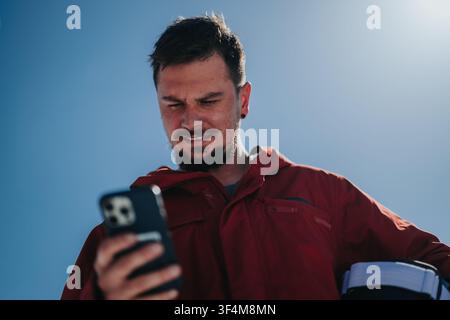 Young man in red jacket checking smartphone outdoors while holding a helmet under his arm Stock Photo