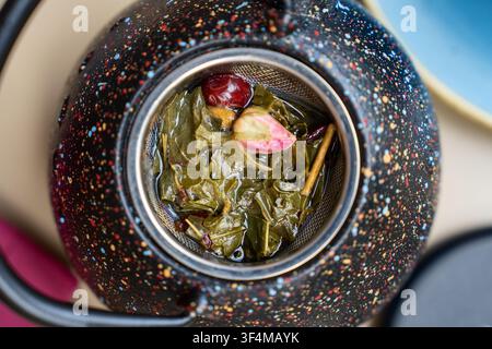 Fragrant botanical tea blend with dried foliage, colorful fruits and blooming rosebud inside a metal strainer within a speckled porcelain kettle Stock Photo