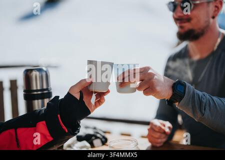 Two friends toasting hot drinks in disposable cups beside a thermos outdoors on a snowy day Stock Photo