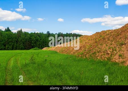 Sandy quarry boundary with large soil heaps invading a lush green meadow near a forest, reflecting industrial landscape changes and environmental impa Stock Photo