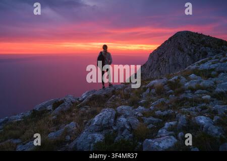 Woman with backpack standing on rocky mountain top at sunset Stock Photo