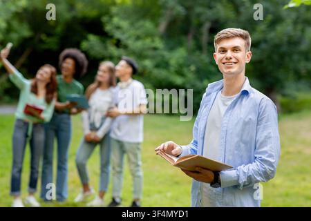 Education First. Student Guy Posing With Book While His Classmates Taking Selfie Stock Photo