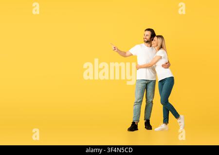 Happy Couple Embracing And Pointing Together, Yellow Background Stock Photo