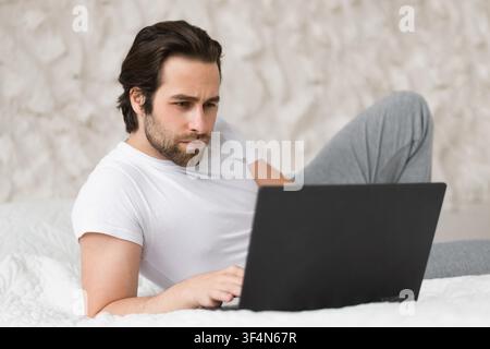 Focused Caucasian Man Working On Laptop While Lying On Bed, At Home Office Concept Stock Photo