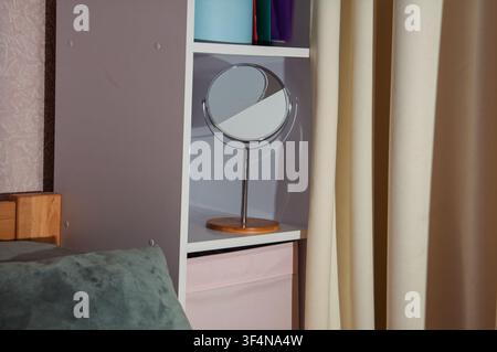 This close-up shot captures a sleek, round vanity mirror with a metal stand and wooden base, placed on a white shelf beside a window draped with light Stock Photo