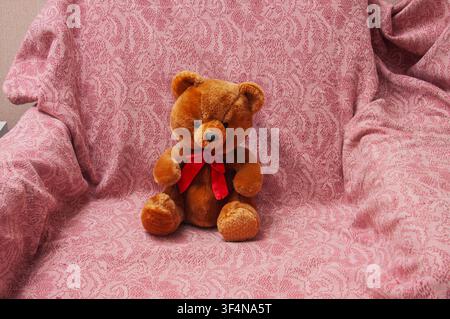 A fluffy brown teddy bear wearing a red bow tie is seated on a textured pink blanket with an intricate floral pattern Stock Photo