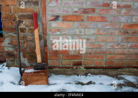 A sledgehammer and crowbar lean against a rough brick wall, with patches of snow and dirt at the base Stock Photo