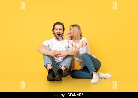 Happy Young Man And Woman Laughing Together Sitting On Floor, Yellow Background Stock Photo