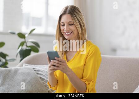Smiling Young Woman Using Smartphone At Home, Relaxing On Sofa Stock Photo