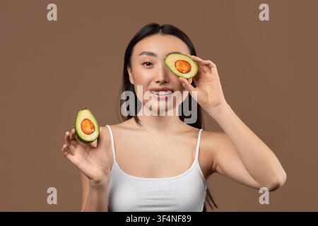 Smiling Asian Woman Holding Avocado Halves On Brown Background, Healthy Eating Concept Stock Photo