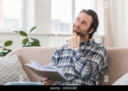 Thoughtful Young Caucasian Man Reading Book At Home, Relaxing On Couch Stock Photo