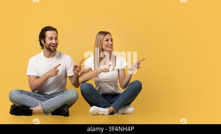 Happy Man And Woman Pointing While Sitting Together On Yellow Background Stock Photo